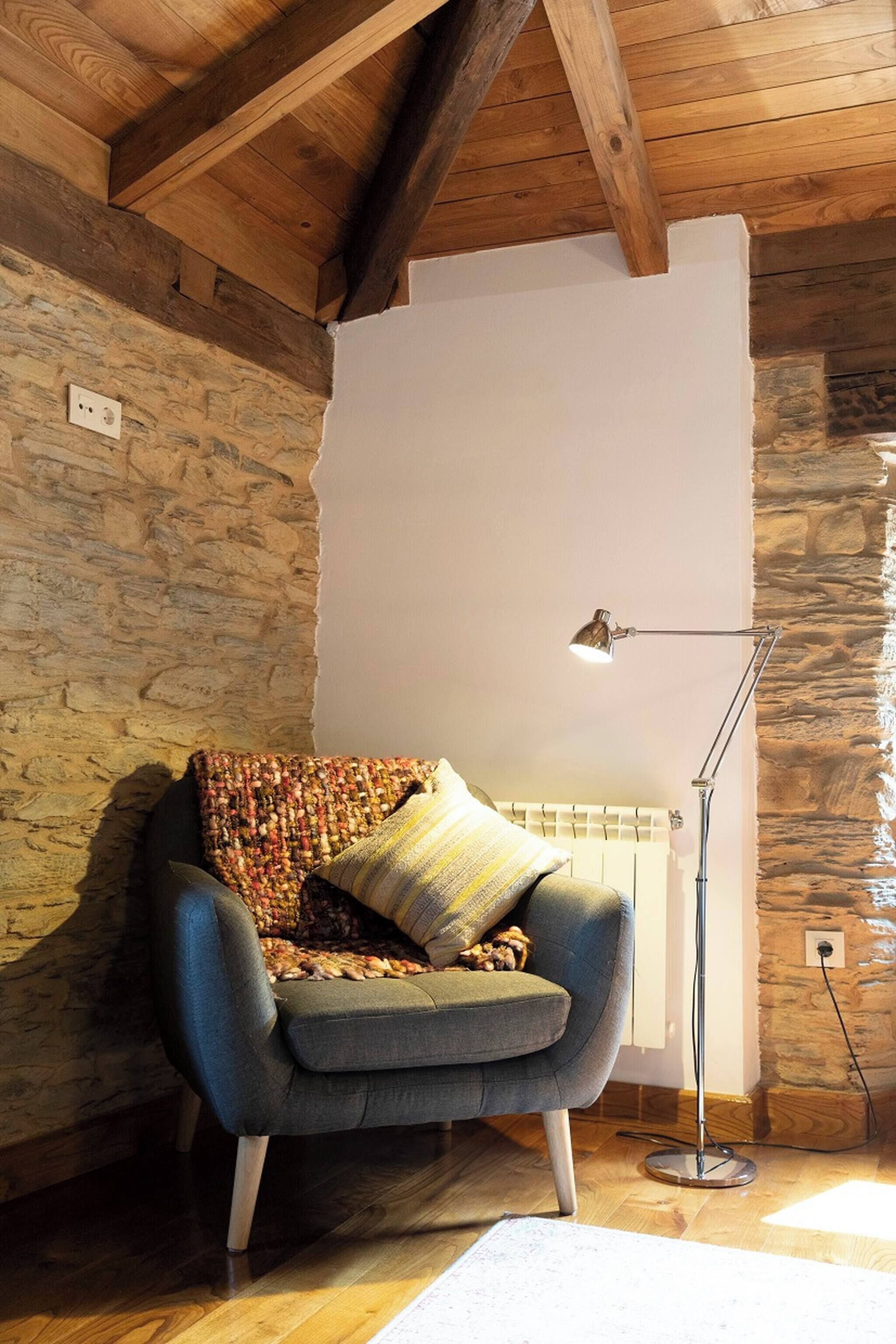 A cozy room with a stone wall, a wooden ceiling, a blue armchair, a floor lamp, and a radiator.