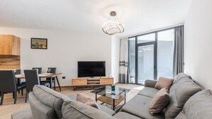 A modern living room with a large grey sofa, a glass coffee table, a flat-screen TV, and a dining area with a wooden table and chairs. - Hill Side Suite 1 by PS (Durrës)