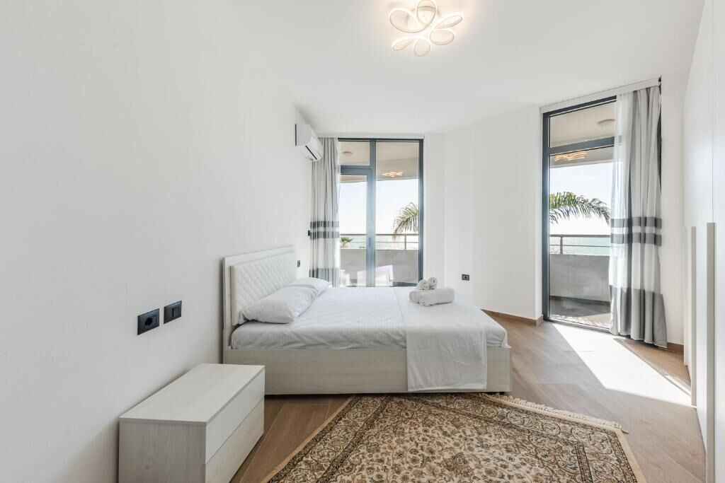 Quarto moderno com uma cama grande, uma mesa de cabeceira, um tapete estampado e uma varanda com cortinas.
