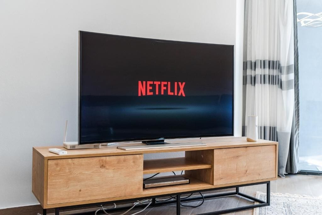 A modern living room with a large flat-screen TV on a wooden TV stand, a white remote, and a small white speaker.