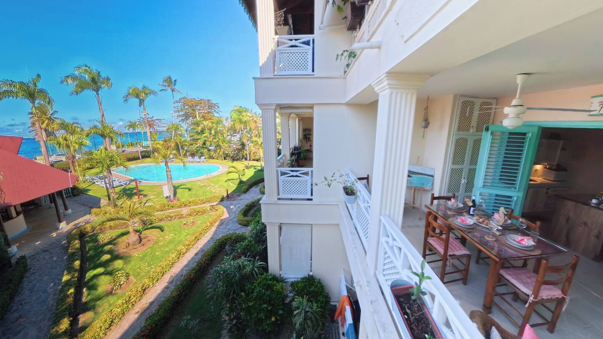 Uma varanda com vista para uma piscina, vegetação exuberante e uma área de jantar com cadeiras e mesa.