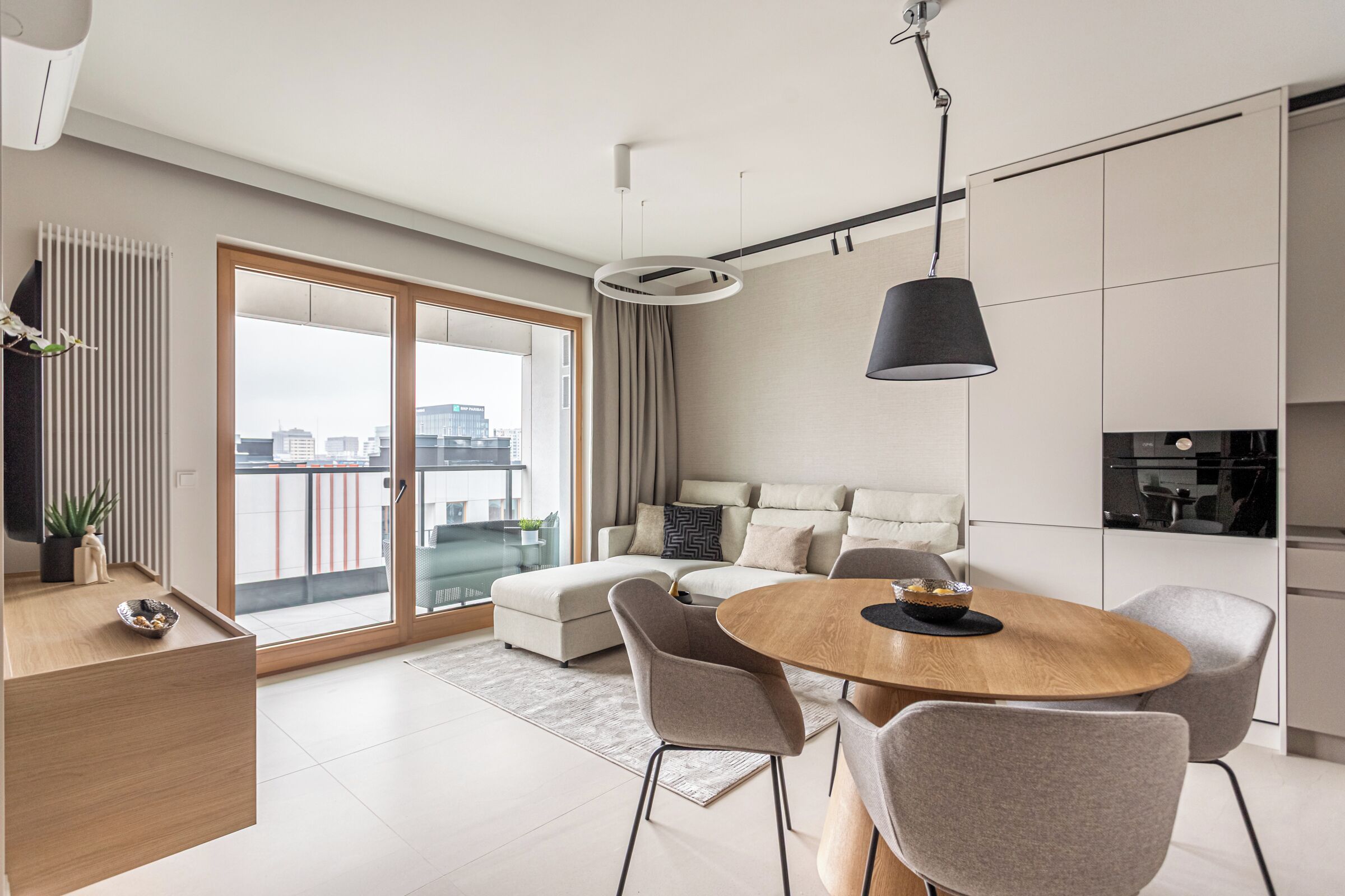 Ein modernes Wohnzimmer mit einer Couch, einem Esstisch und Stühlen. Ein großes Fenster bietet Blick auf Gebäude.