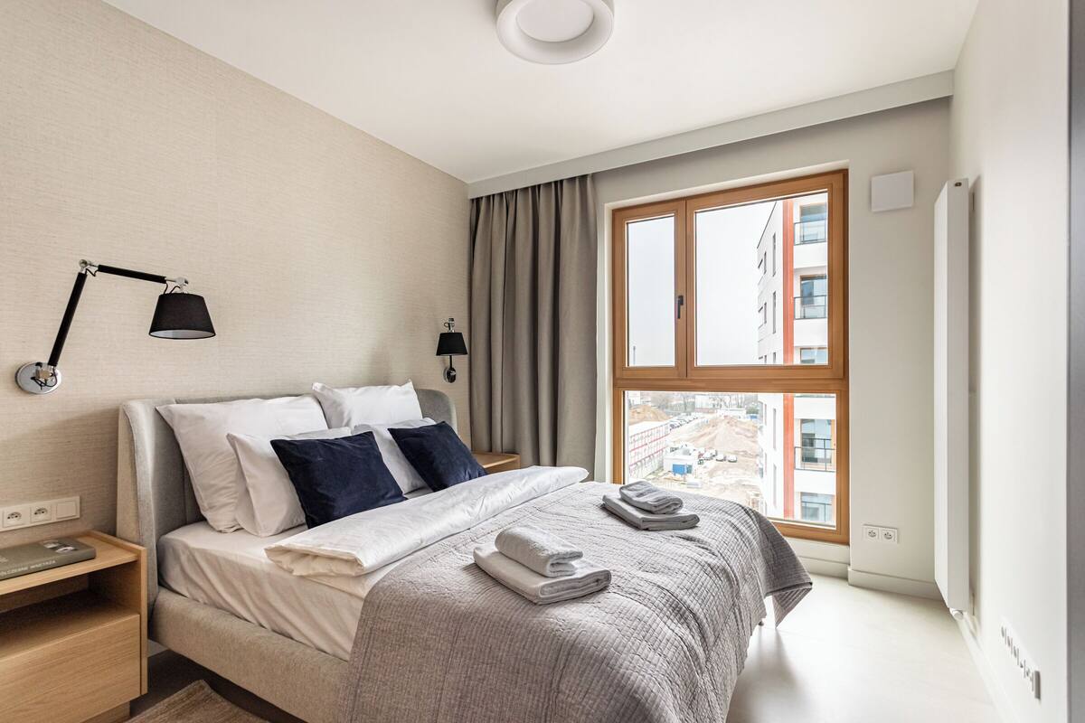 Egy modern hálószoba, amelyben egy nagy ágy, két éjjézőlámpa, ablakfüggönnyel ellátott ablak található, és az ablakon keresztül a környező épületekre nyílik kilátás.