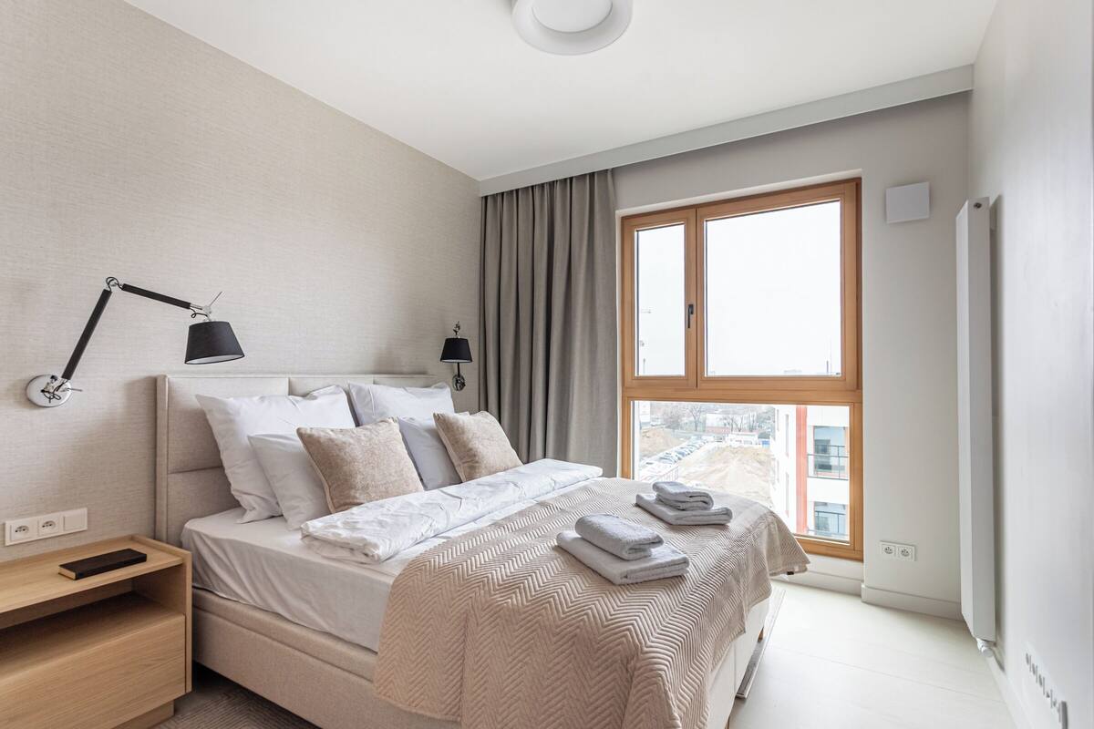 Egy modern hálószoba, melyben egy nagy ágy, éjjeli lámpák, egy éjjeliszemély és egy nagy ablakon át nyíló kilátás a városra található.