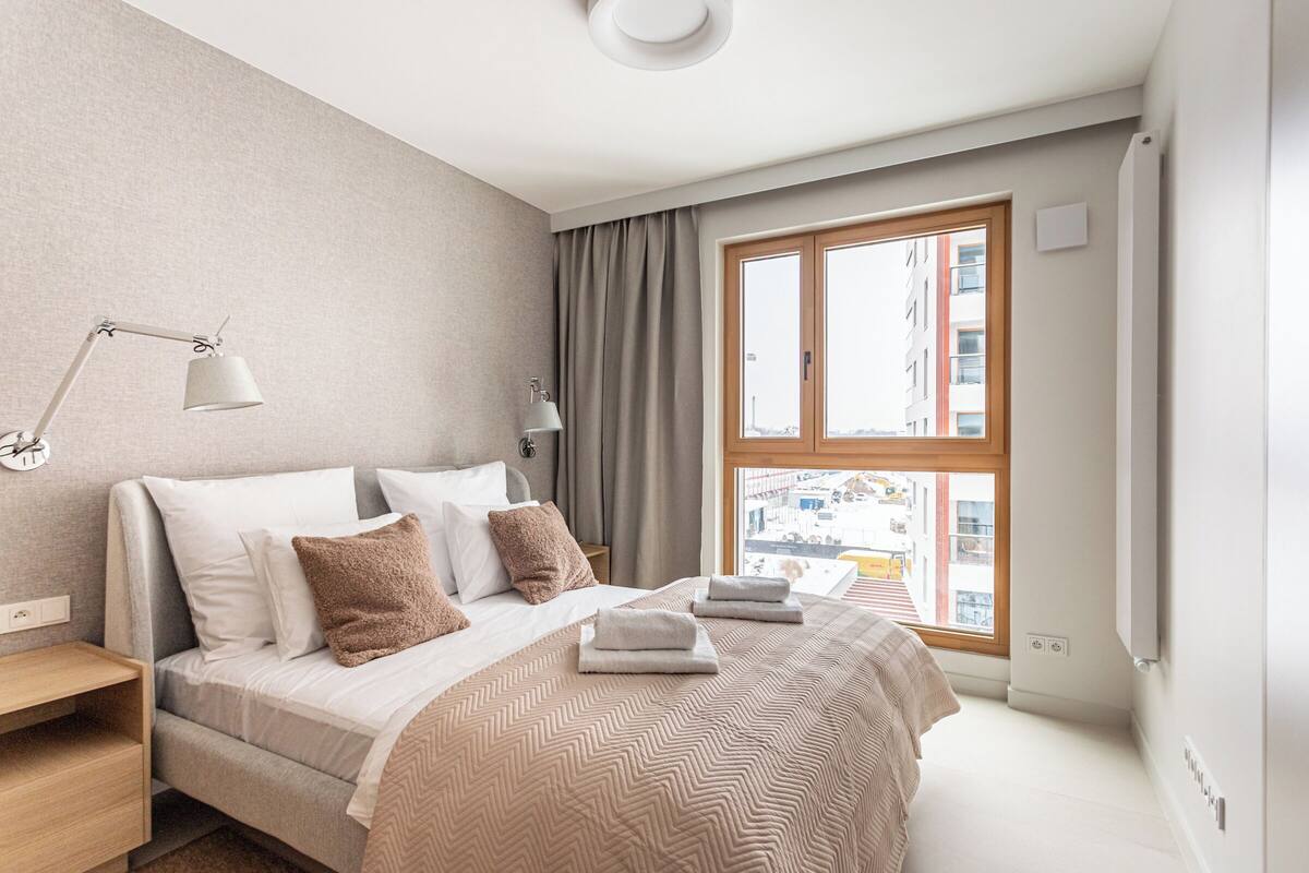 Egy modern hálószoba, amelyben található egy ágy, egy éjjeli lámpa és egy nagy ablak, melyből a városra nyílik kilátás.