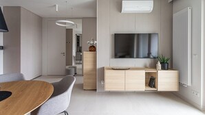 Ένα μοντέρνο σαλόνι με ένα ξύλινο τραπέζι τραπεζαρίας, γκρι καναπέδες, μια τηλεόραση επίπεδης οθόνης τοποθετημένη στον τοίχο, μια εντοιχισμένη ξύλινη ντουλάπα και ένα καλοριφέρ.