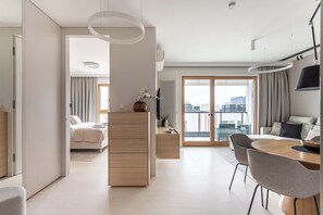 Une chambre d’hôtel moderne comprenant un lit, une commode en bois, un coin repas avec une table et des chaises, et une grande fenêtre donnant sur la ville.