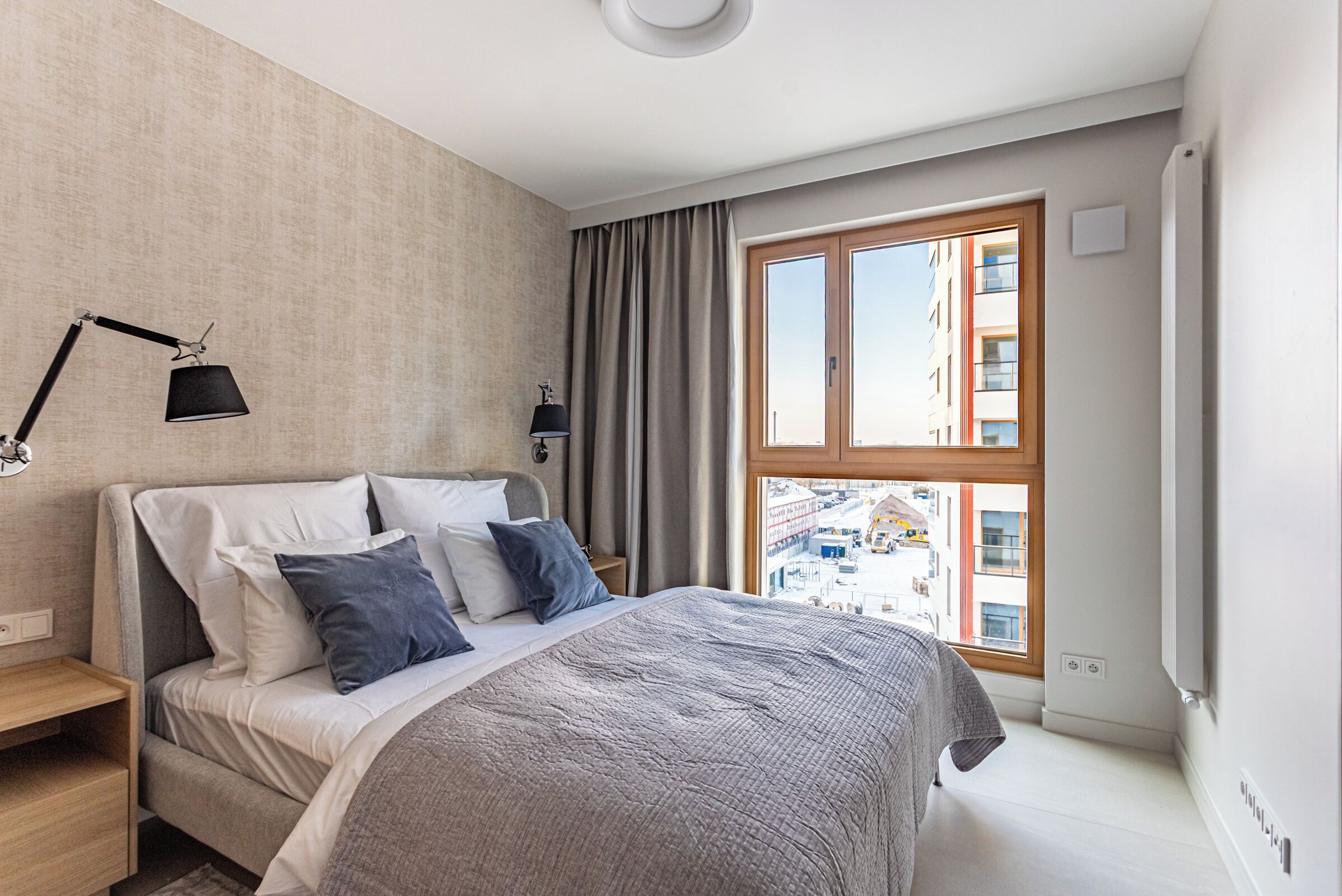 Une chambre à coucher moderne avec une grande fenêtre, un lit avec une literie grise, une table de chevet et une lampe.