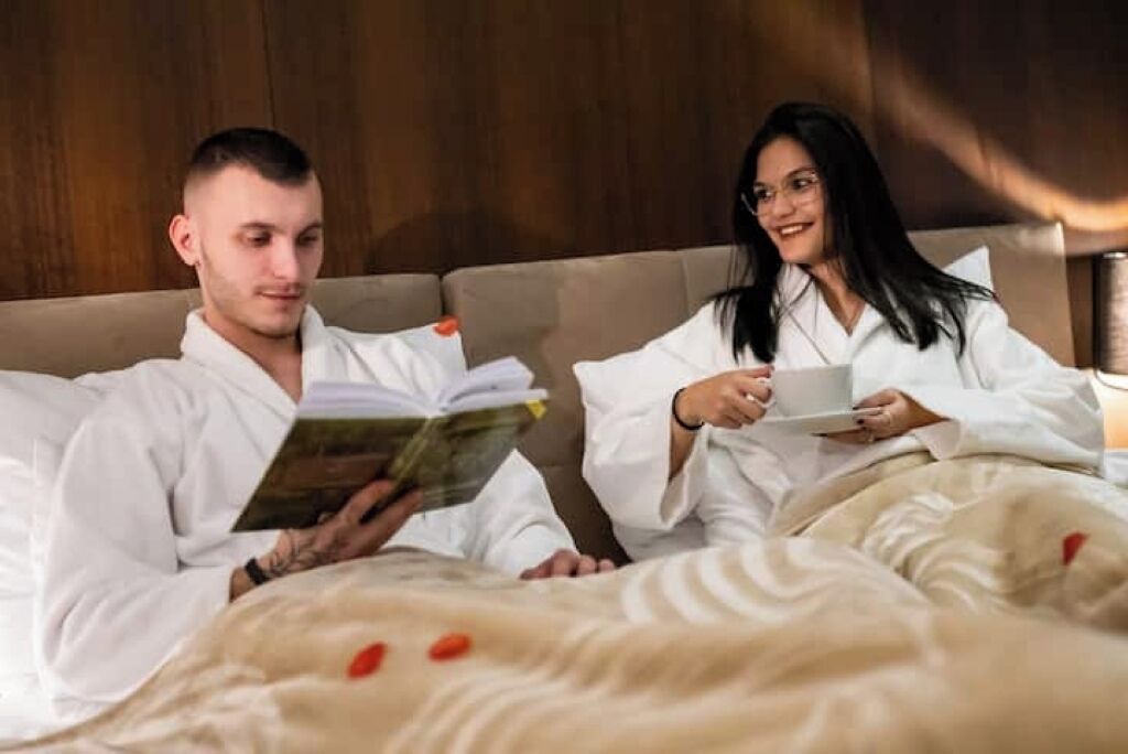 A couple in bed with a book and a cup, in a room with wooden headboard and beige bedding.
