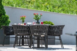 An outdoor dining area with wicker chairs and a round table, set against a grey wall with greenery in the background.