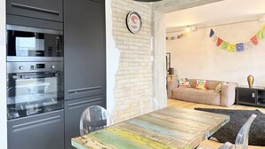 Una cocina con microondas, horno y una mesa de comedor con sillas. Hay una pared de ladrillo, una lámpara colgante y un sofá con cojines de colores en la sala de estar.