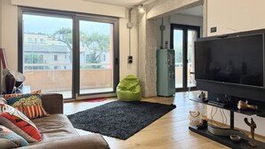 Una sala de estar con un televisor grande, un sillón orejero verde, una alfombra negra y vistas de edificios.