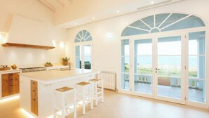 A spacious kitchen with a central island, wooden cabinets, and a large window with a view of the sea. - Veles Dos – Seafront Villa with Private Pool and Stunning Views (Sa Marina - Barcarés - Manresa)