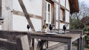 A rustic outdoor dining table set with wine glasses, plates, and a bottle, positioned in front of a building with exposed wooden beams and a window. - Remark Studios: Historic and modern Apartment (Burgwedel)
