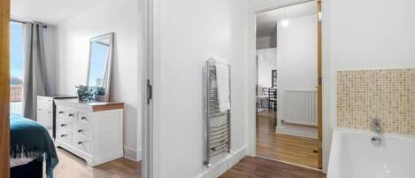 Un dormitorio moderno con cama, cómoda y un espejo grande. Se ve un pasillo que conduce a un baño con bañera y zona de ducha con azulejos.