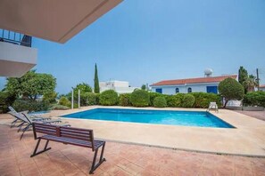 A swimming pool with lounge chairs and a bench, surrounded by greenery and buildings in the background. - Modern stylish 2 bedroom apartment with communal pool in Neo Chorio Village (Neyohoryo Baf)