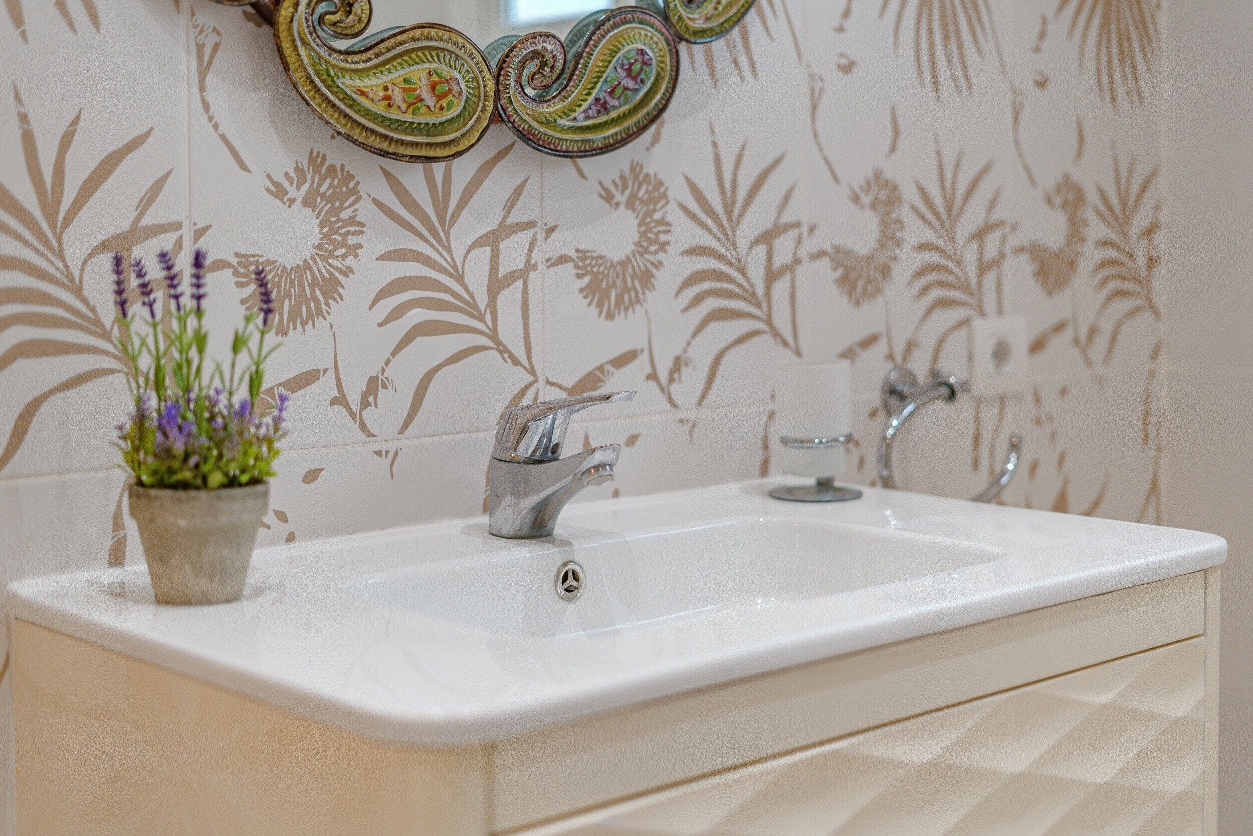 A bathroom with a white sink, two faucets, a mirror with a decorative frame, and a floral wallpaper pattern.