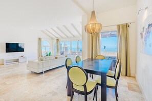 A spacious living area with a large dining table, comfortable seating, a flat-screen TV, and a view of the sea.