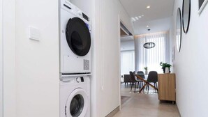 Een moderne keuken met een ingebouwde wasmachine, een eethoek met een houten tafel en stoelen, en uitzicht op de woonkamer door een open deur.