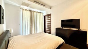A hotel room with a bed, a television on a cabinet, a window with curtains, and a wall-mounted air vent. - Fort Cambridge, Sliema 1 BDR (Sliema)