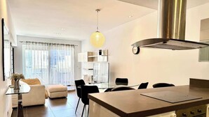 A modern kitchen with a central island, a dining area with a glass table, and a living space with a sofa and chairs. - Fort Cambridge, Sliema 1 BDR (Sliema)