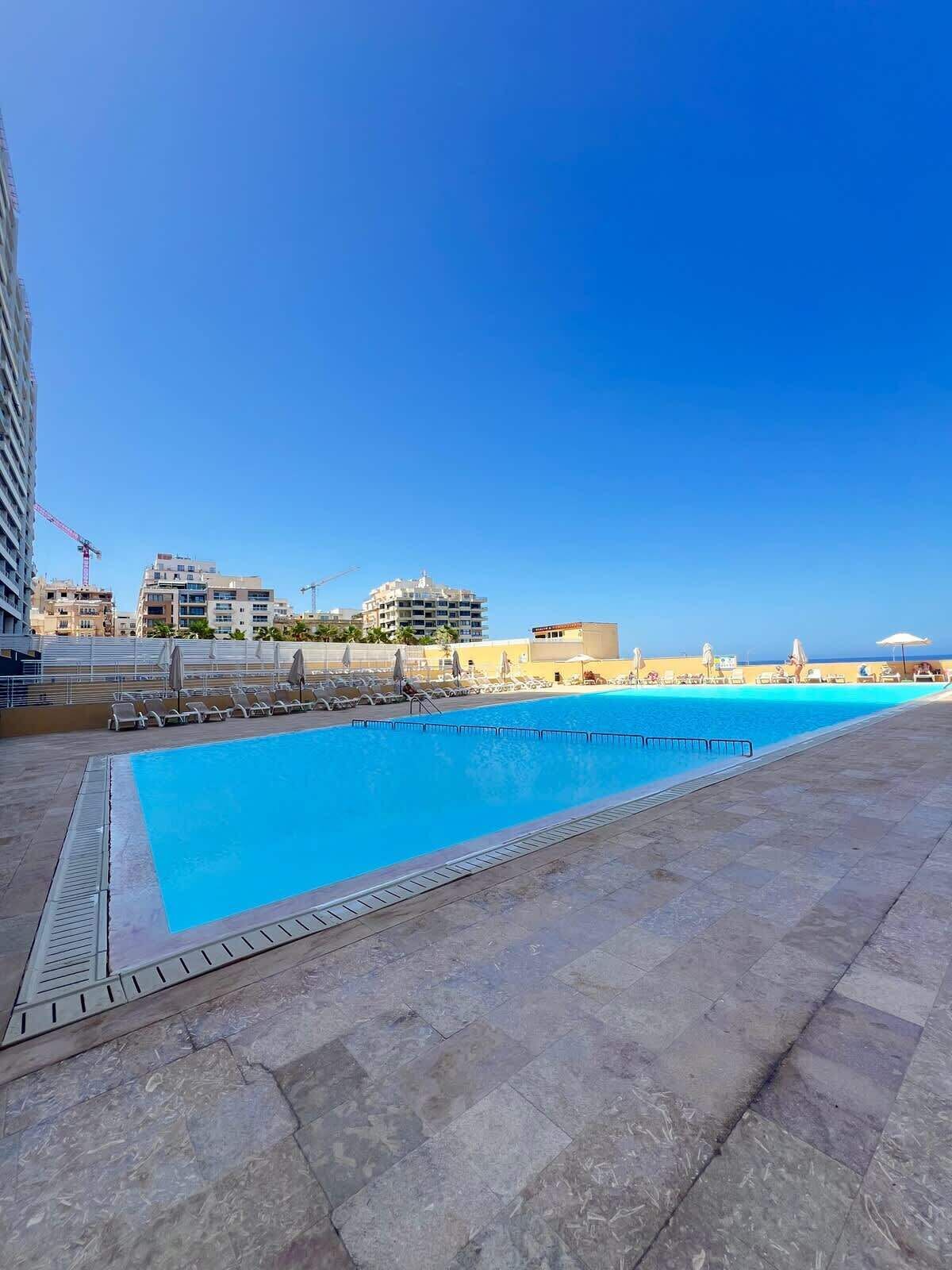 A swimming pool with lounge chairs and umbrellas, surrounded by buildings and a clear sky.
