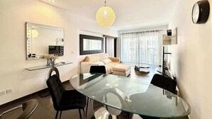 A modern living room with a glass dining table, black chairs, a beige sofa, a large mirror, and a hanging light fixture. - Fort Cambridge, Sliema 1 BDR (Sliema)