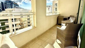 A balcony with a sofa, wicker chairs, and a view of buildings and a construction crane. - Fort Cambridge, Sliema 1 BDR (Sliema)