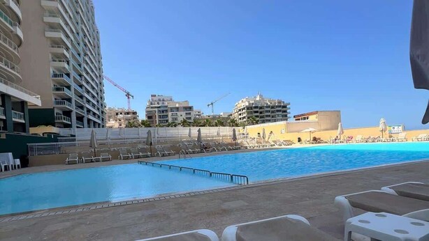A poolside area with lounge chairs, a swimming pool, and a view of a high-rise building and other structures in the background. - Fort Cambridge, Sliema 1 BDR (Sliema)