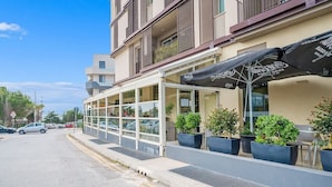 Ein Gebäude mit Glasfassade, einer Außenbestuhlung mit Sonnenschirmen und Blumentöpfen.