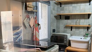 Une cuisine compacte équipée d’un four à micro-ondes, d’un grille-pain et d’un évier. On y trouve des étagères en bois au-dessus du plan de travail et une fenêtre avec des rideaux.