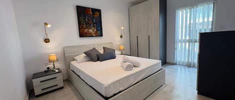A modern bedroom with a large bed, bedside tables, a wardrobe, and a window with blinds.