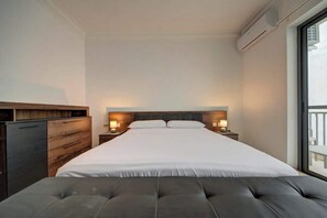 Ein modernes Hotelzimmer mit einem großen Bett, Nachttischlampen, einem hölzernen Kopfteil, einer grauen Sitzbank und einem großen Fenster mit Balkonblick.