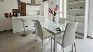 Et moderne køkken med hvide skabe, en central ø og et spiseområde med et glasbord og hvide stole.