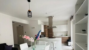 Et moderne køkken med hvide skabe, en central ø og et spiseområde med et glasbord og hvide stole.