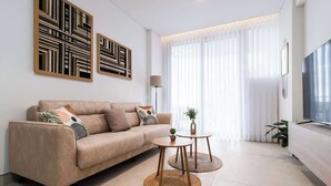 A modern living room with a beige sofa, two round wooden tables, and a large TV on a white cabinet. - Shoreline Residence Brand NEW 1 BDR Apartment (Kalkara)