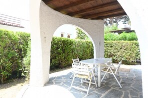 Une terrasse extérieure avec un sol en pierre, des meubles blancs et une vue dégagée sur l’extérieur, accessible par une arche.