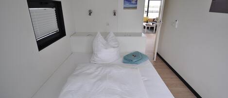 A single bed with a white mattress and pillows, a small window with blinds, and a blue towel on the side.