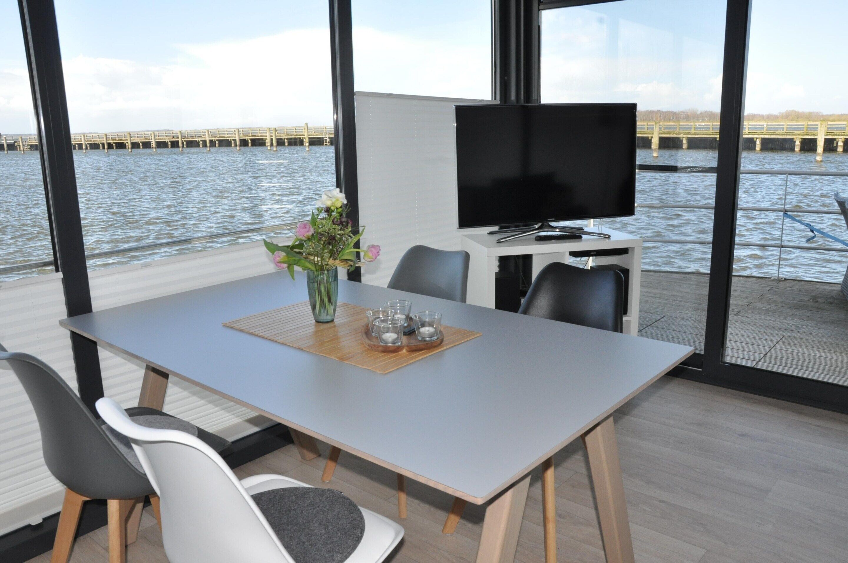 A modern dining area with a large table, chairs, a flat-screen TV, and a view of a bridge over water.