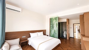 Habitación de hotel con cabecera de madera, cama con ropa blanca, mesita de noche, aire acondicionado instalado en la pared y vista al pasillo.