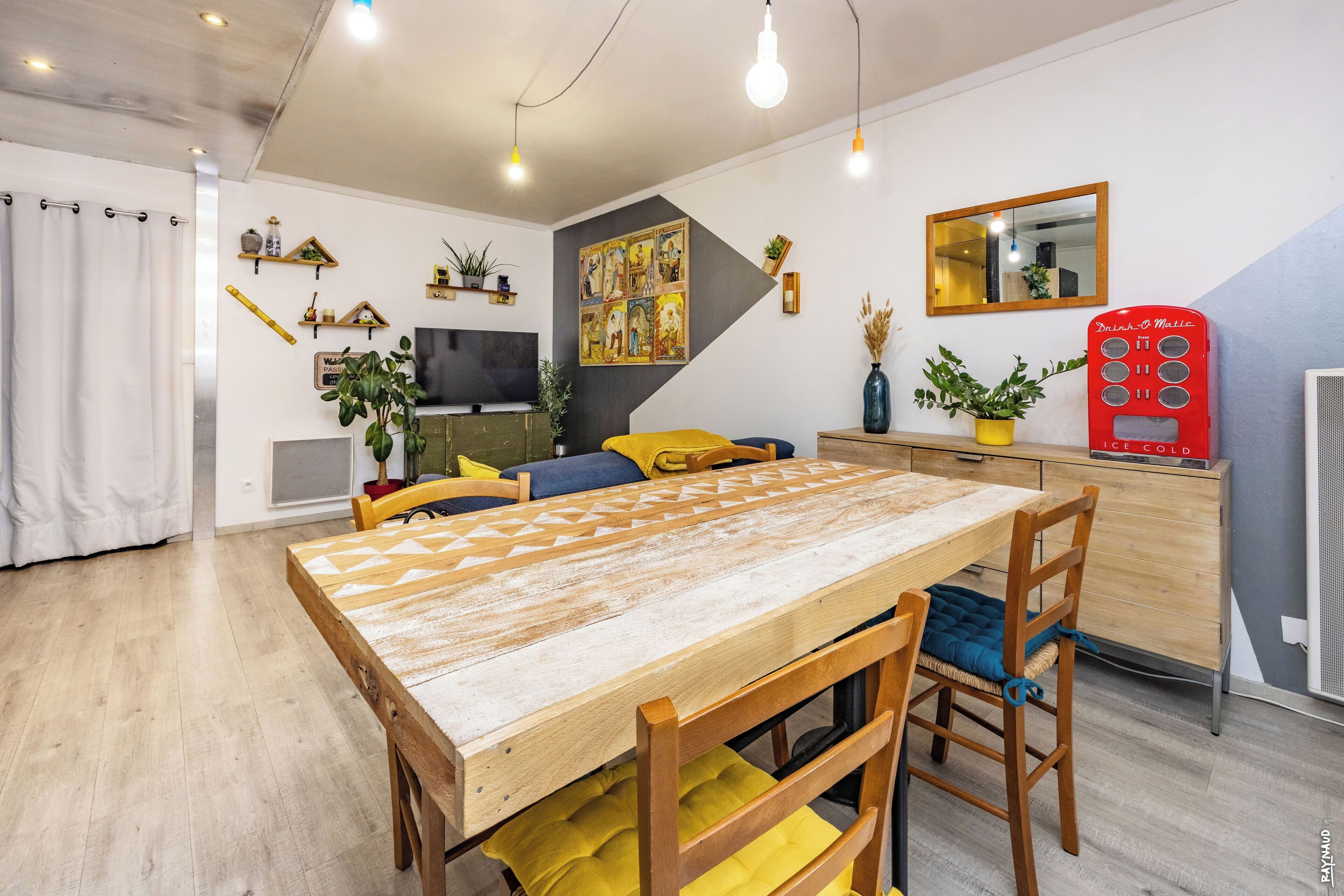 A modern living room with a wooden dining table, yellow cushions, and a red ice cream vending machine.