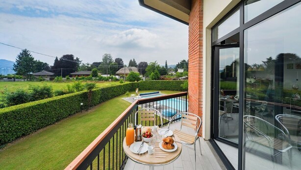 Een balkon met een ontbijttafel, een glazen deur naar een zwembad en uitzicht op een verzorgde tuin en omliggende huizen.