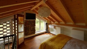 Een slaapkamer met een schuine balkenlaag, een stenen muur, een bed met een gele deken, een televisie aan de muur en een groot raam.