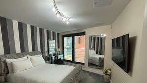 A bedroom with a large bed, a TV mounted on the wall, a desk with a chair, and a window with blinds.