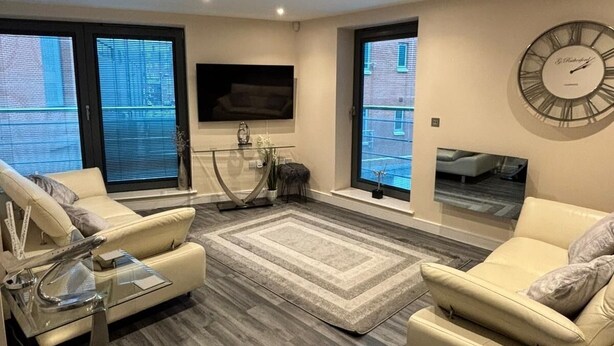 A modern living room with two leather sofas, a glass coffee table, a large wall clock, and a flat-screen TV mounted on the wall.