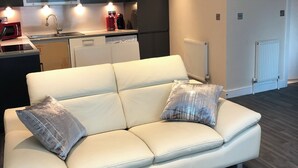 A modern living room with a white leather sofa, two decorative pillows, a glass coffee table, and a kitchen area in the background.