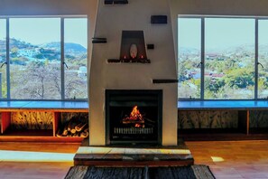 A fireplace with a fire, large windows, and a view of a hilly landscape. - Stunning Panoramic View Swimming Pool 3-bedroom Apartment in the Luxury of Eros. (Windhoek)