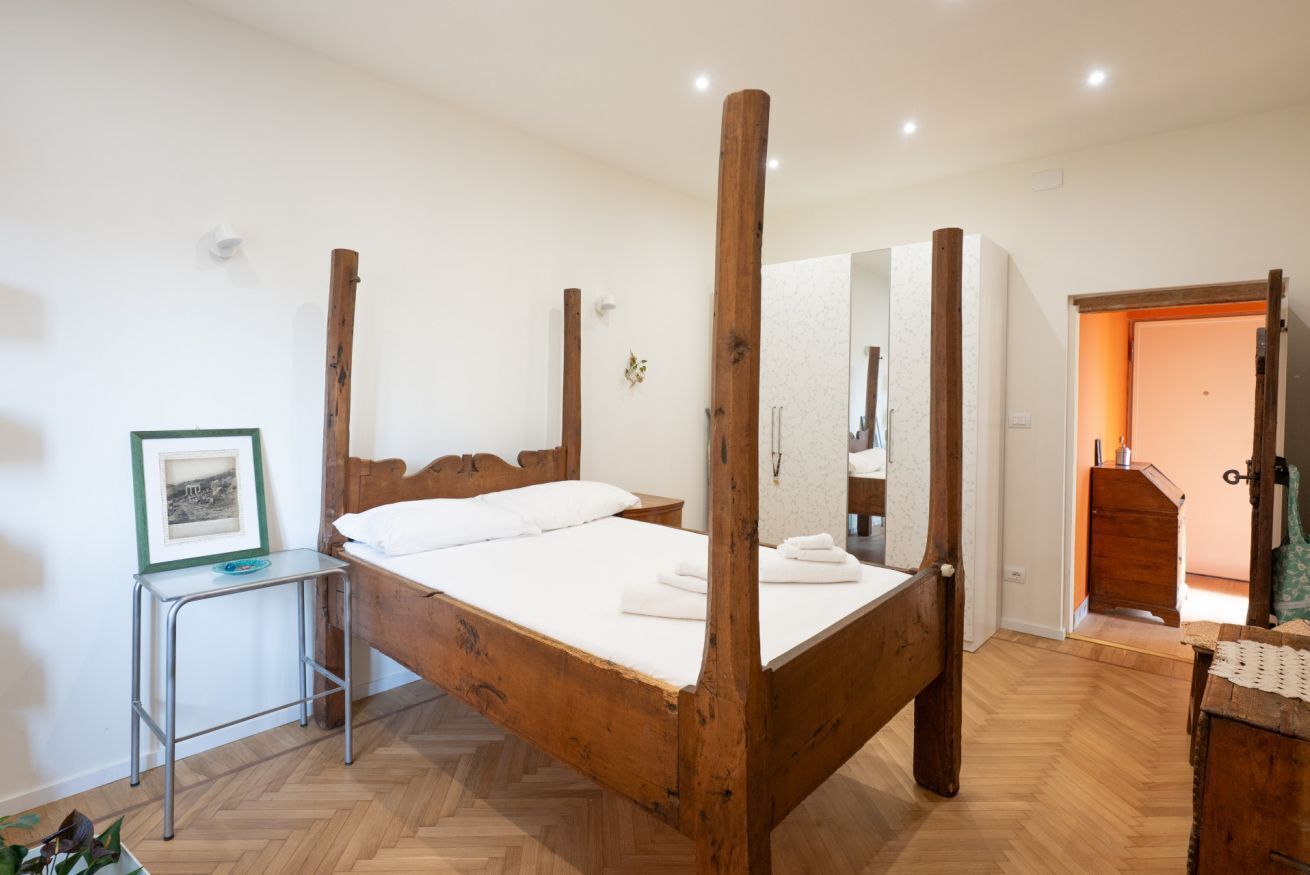 A wooden four-poster bed with a white mattress, a small metal side table, and a framed picture on the wall.