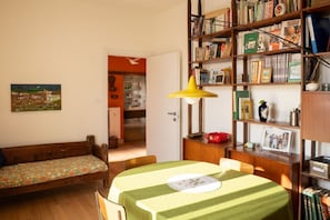 Una habitación con un banco de madera, una mesa cubierta con mantel verde, una estantería llena de libros y un cuadro en la pared.