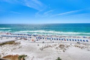 Ein Strand mit Reihen blauer Sonnenschirme und Liegen, ein sandiger Uferbereich und das Meer im Hintergrund.